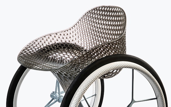 View of the middle section of a geometrical, 3D-printed chair seat from a 3D-printed prototype of a customized, futuristic-looking wheelchair, using multiple 3D printing materials. The seat is lattice-structured and made of a translucent gray resin. The wheel spokes are made of 3D-printed metal.