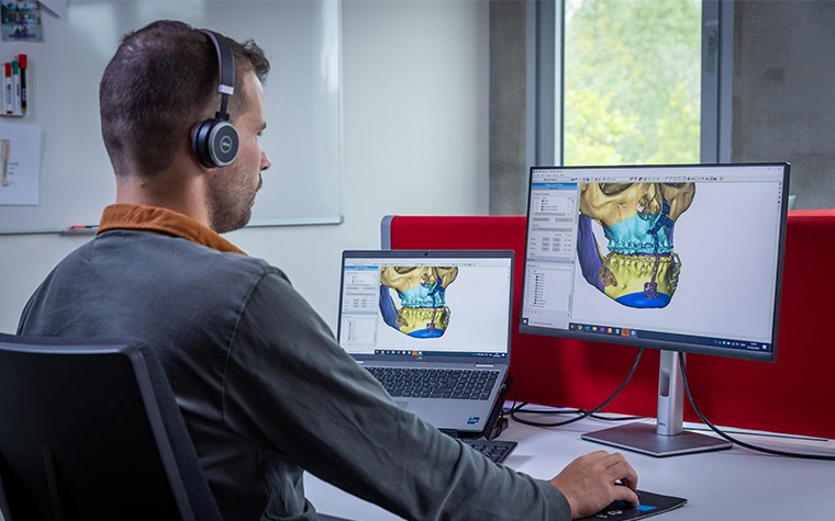 Ein Mann mit Kopfhörern schaut auf zwei Computerbildschirme mit Materialise-Software, auf denen jeweils ein Totenkopf zu sehen ist.