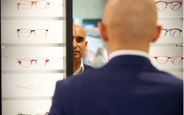 Man looking into a mirror surrounded by eyeglasses on shelves