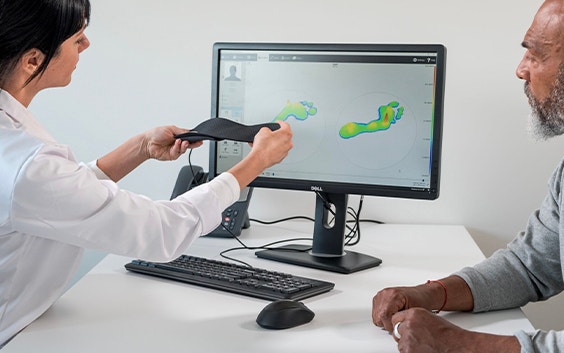 Doctor holding up a personalized insole while talking to a patient and looking at a screen with footscan software pulled up
