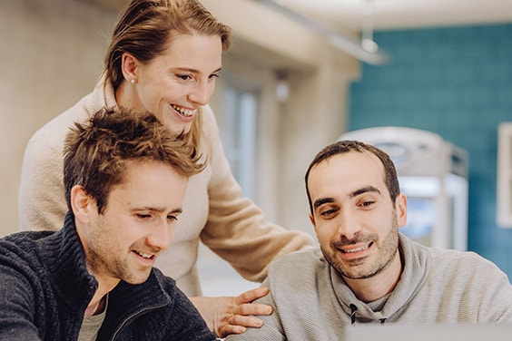 Two men sitting and smiling with a woman standing behind them smiling, all looking downwards