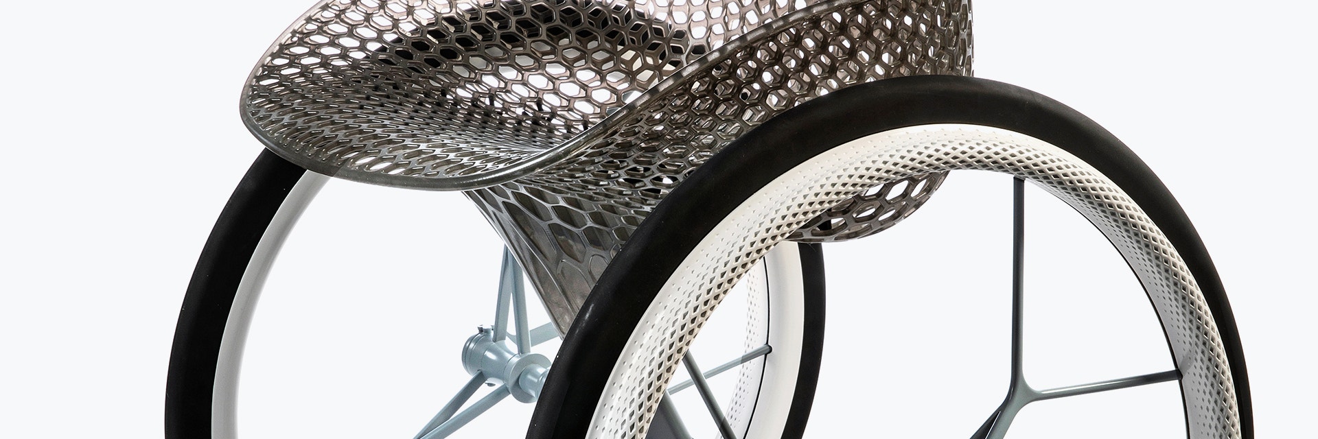 View of the middle section of a geometrical, 3D-printed chair seat from a 3D-printed prototype of a customized, futuristic-looking wheelchair, using multiple 3D printing materials. The seat is lattice-structured and made of a translucent gray resin. The wheel spokes are made of 3D-printed metal.