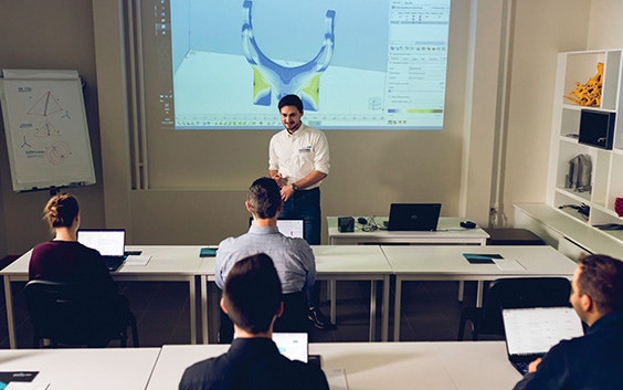 Eine Lehrkraft unterrichtet ein Klassenzimmer voller Studenten vor einem Bildschirm mit einem 3D-Design