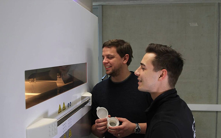 Two men look through a window on a laser sintering printer to observe parts being 3D printed