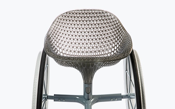 Frontal view of a 3D-printed prototype of a customized, futuristic-looking wheelchair, using multiple 3D printing materials. The seat is lattice-structured and made of a translucent gray resin. The foot-rest and wheel spokes are made of 3D-printed metal.