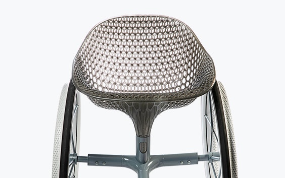 Frontal view of a 3D-printed prototype of a customized, futuristic-looking wheelchair, using multiple 3D printing materials. The seat is lattice-structured and made of a translucent gray resin. The foot-rest and wheel spokes are made of 3D-printed metal. 