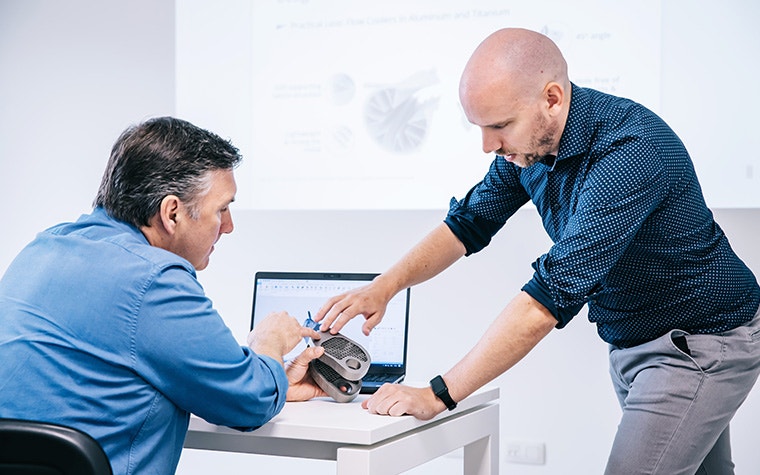 A Materialise Academy trainer and trainee looking at a metal 3D-printed part together in front of a laptop and projector showing the design in Materialise Magics