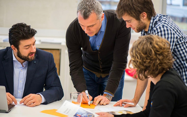 Group of people around a table, looking at a man writing with a marker