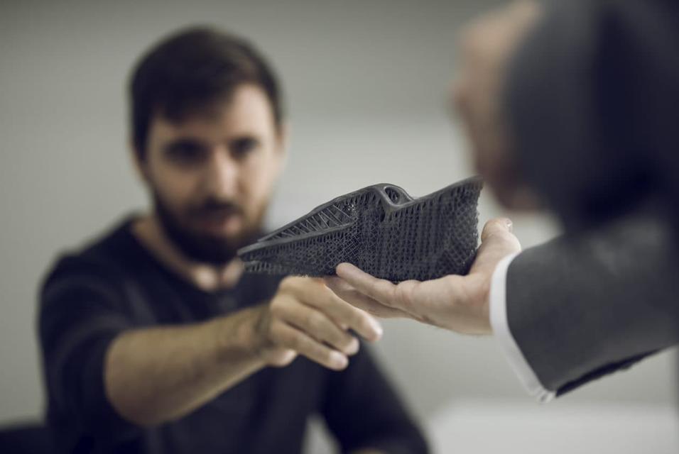 Man in the blurred background pointing to a 3D-printed part with support structures that's held up by another person