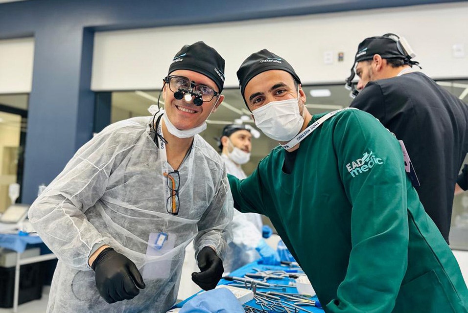 Two medical professionals wearing surgical attire, masks, and magnifying loupes posing together during a hands-on Materialise CMF Wet Lab training session, with instruments and participants in the background.
