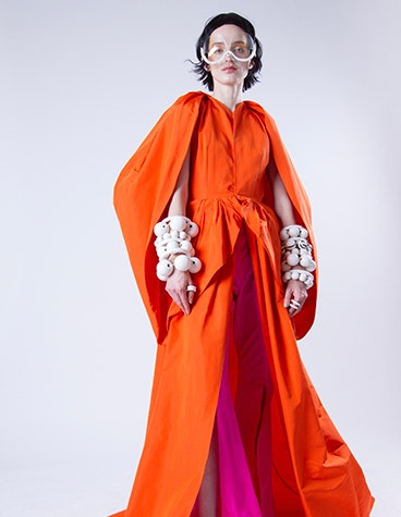 Model trägt ein orangefarbenes Kleid, klobige Armbänder und eine weiße Sonnenbrille, entworfen von David Ring