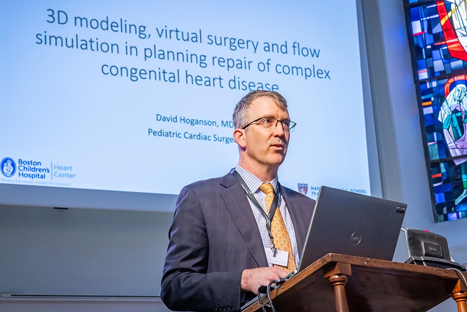 Pediatric Cardiac Surgeon Dr. David Hoganson standing with a laptop on a lecturn in front of a whiteboard, giving a presentation on 3D modeling, virtual surgery, and flow simulation in planning repair of complex congenital heart disease at Materialise's 3D Planning and Printing in Hospitals Forum (2025).