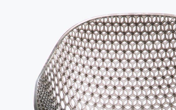 Close-up view of a geometrical, 3D-printed chair seat from a 3D-printed prototype of a customized, futuristic-looking wheelchair, using multiple 3D printing materials. The seat is lattice-structured and made of a translucent gray resin.
