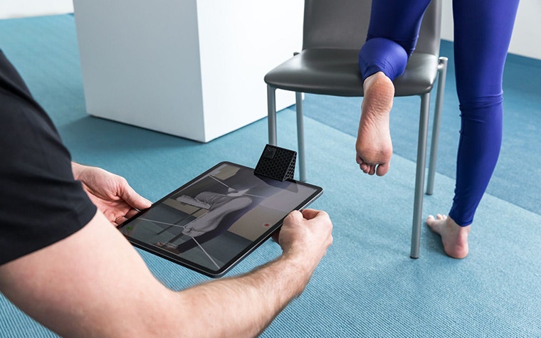 A patient kneels on a chair while a doctor looks over the 3D scan of her foot on an iPad.