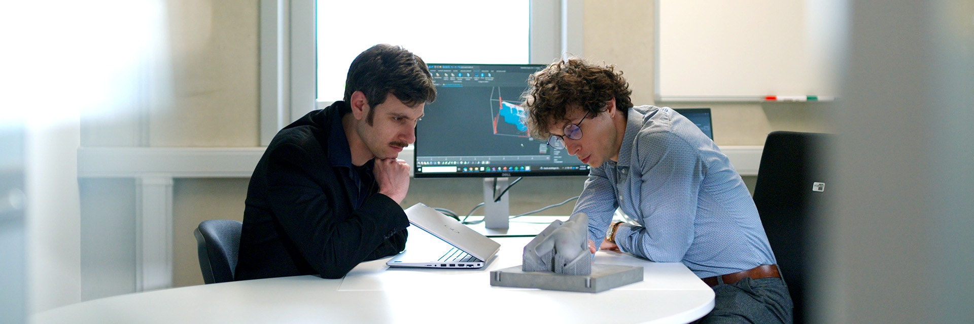 Two men sitting at a desk looking at metal 3D-printed part with thin support structures.