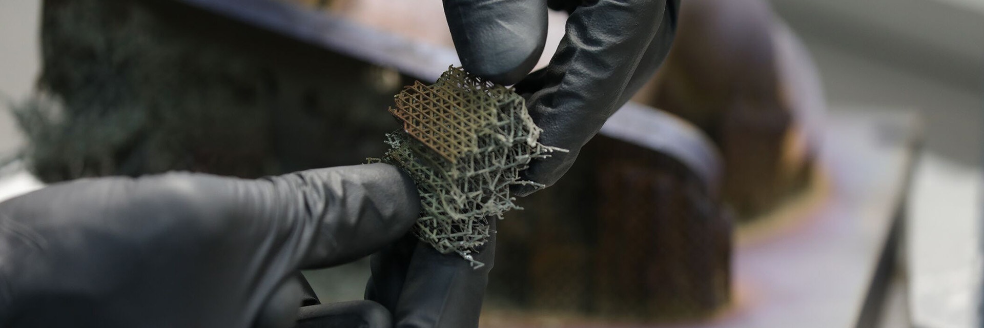 A person with gloves holding a small metal 3D-printed square with many support structures attached after post-processing.