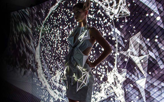 Model posing in front of geometrical images on a screen, wearing a 3D-printed dress