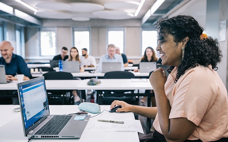 Woman sitting at a computer and smiling in front of a classroom of students on their laptops