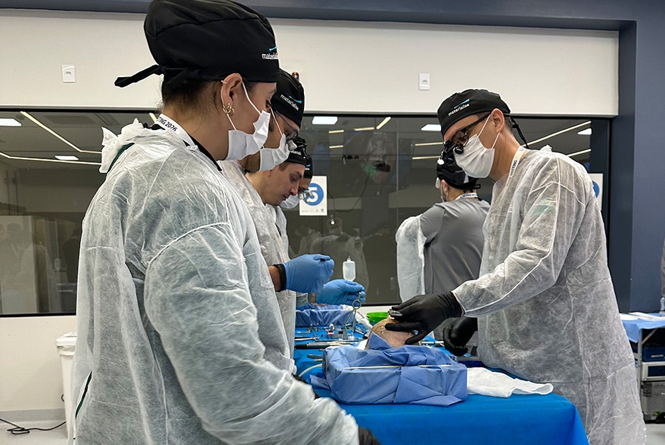Medical professionals wearing protective gowns, masks, and surgical loupes while participating in a hands-on Materialise CMF Wet Lab training session around an operating table.