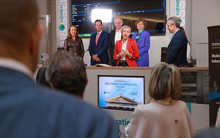 Materialise CEO Brigitte de Vet standing on a podium, speaking to an audience at Euronext Brussels. Members of the executive committee stand behind her, including Director of Corporate Affairs Carla Van Steenberegen, CFO Koen Berges, and founders Fried and Hilde Vancraen. A screen on the podium says 'Euronext Brussels welcomes Materialise to the Market'