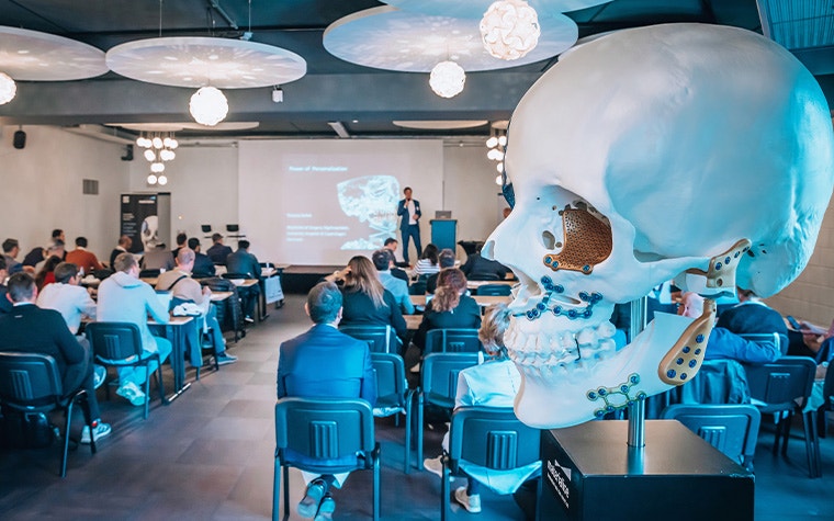 A large skull model with personalized implants attached in a meeting room with a presenter standing in front of a screen and event attendees sitting in seats at tables