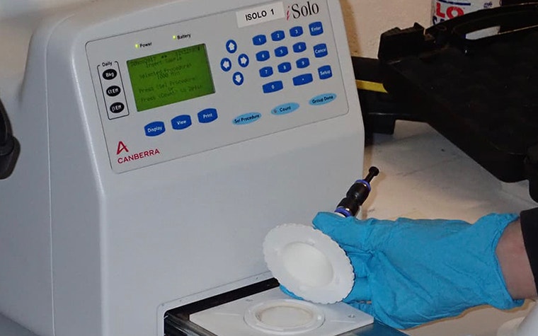 A worker wearing gloves places a sample in the sample preparation unit of an iSolo alpha/beta counter 