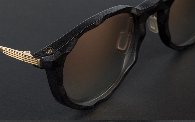 Close-up of the front right corner of black sunglasses from the IMPRESSIO Vortex collection with gold accents