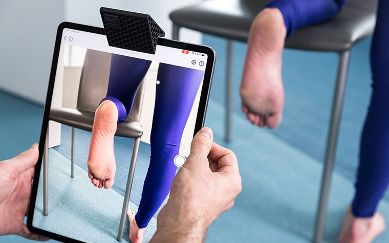 A patient kneels on a chair while a doctor scans the sole of their foot using an iPad and the SAM Mirror attachment.