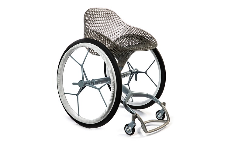 View of a 3D-printed prototype of a customized, futuristic-looking wheelchair, using multiple 3D printing materials. The seat is lattice-structured and made of a translucent gray resin. The foot-rest and wheel spokes are made of 3D-printed metal. 
