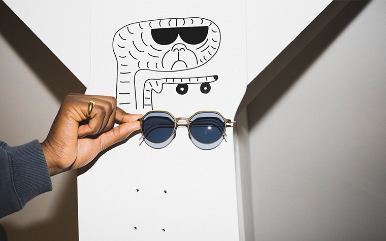 A hand holding a pair of weareannu's 3D-printed Oblique Panto 08 M sunglasses beneath a drawing of a wrinkly, alien-like creature