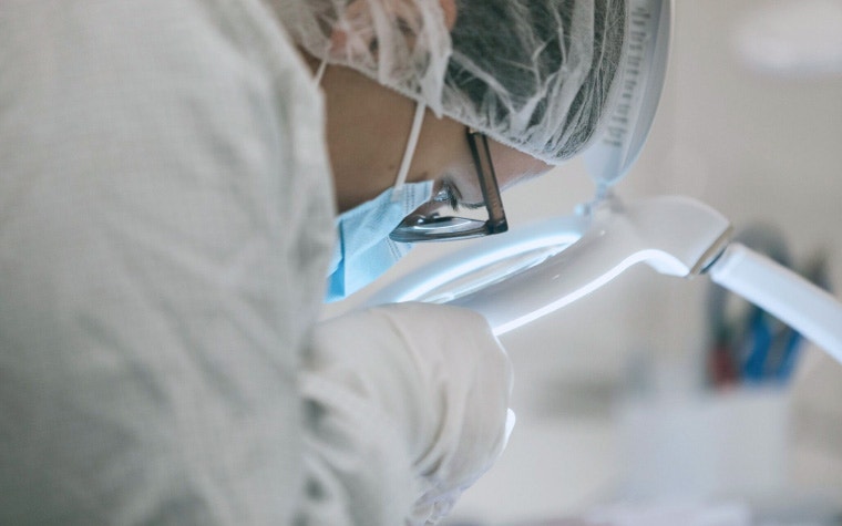 Medical quality engineer wearing protective gear and inspecting a part under a magnifying glass in a cleanroom