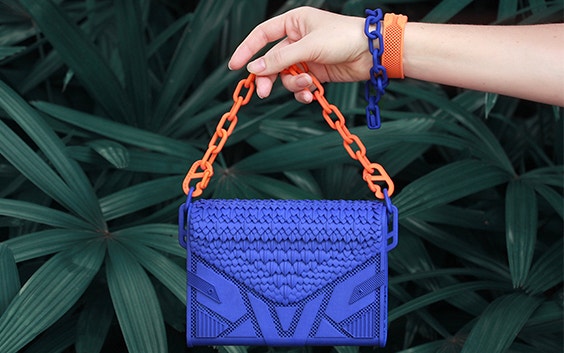 Die Hand hält eine orangefarbene und blaue 3D-gedruckte Handtasche mit Kettengliedern vor Pflanzen