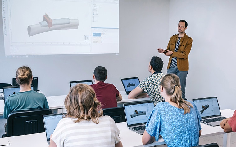 A Materialise Medical expert stands at the front of a classroom full of trainees on their laptops, teaching them about a 3D-printed surgical guide shown in Mimics software on a projector. The trainees are also looking at the same screen on their laptops.