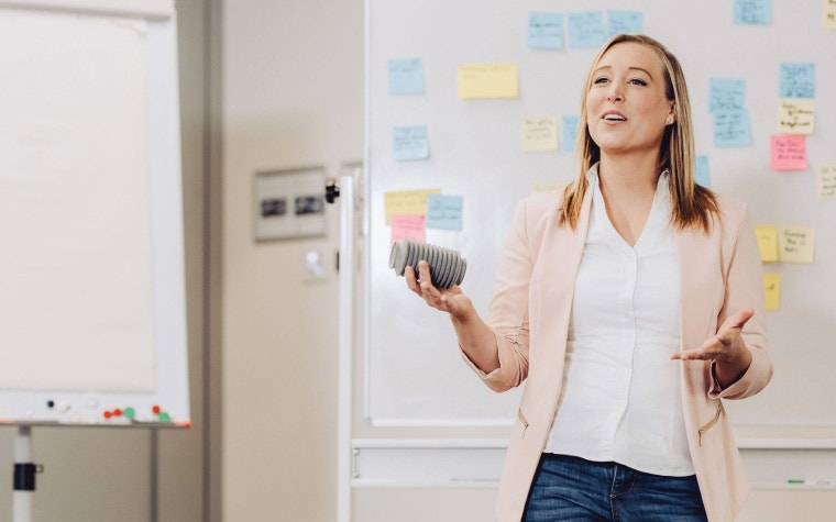 Woman holding a 3D-printed part and speaking in front of a whiteboard with post-it notes attached