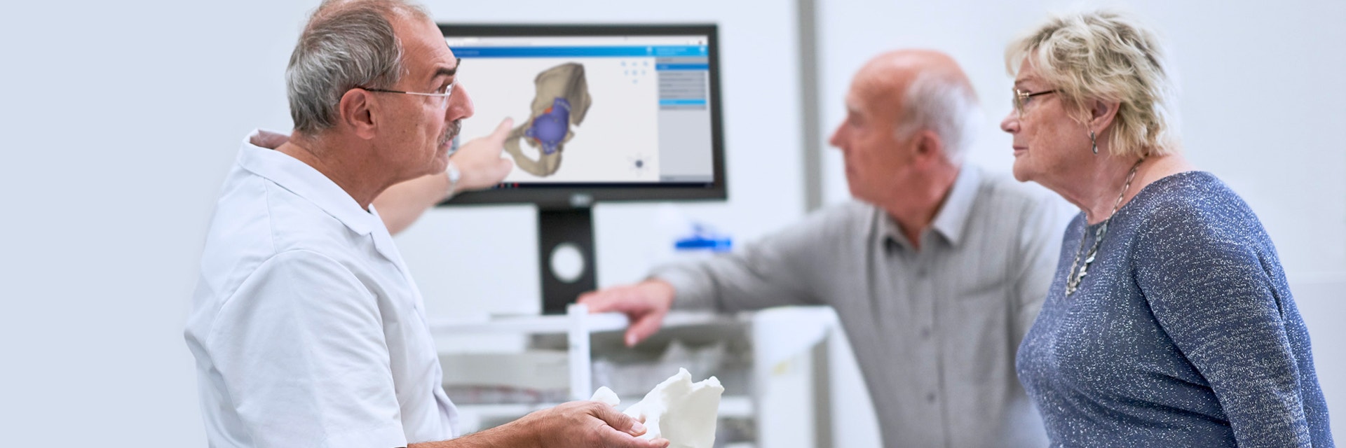 Doctor holding a 3D-printed anatomical model and pointing to a screen including the digital design of a personalized hip implant while talking to a patient