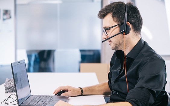 Man wearing a headset and sitting at a desk on his laptop with his hand on a mouse