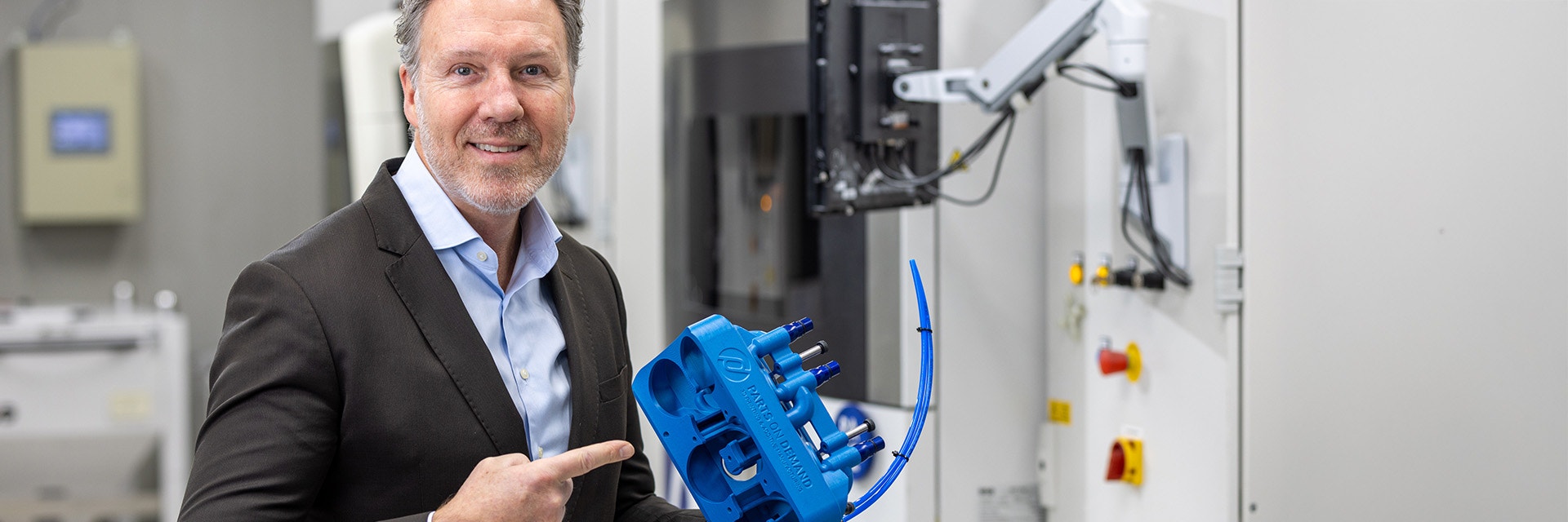 Frank Elbersen, Sales Engineer at Parts on Demand, point to a blue 3D-printed part in front of a 3D printer.