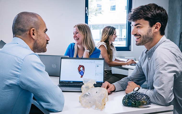 Two men sitting in the foreground, smiling while talking next to a laptop showing an anatomical model and 3D-printed anatomical models. Two women are sitting in the background, facing away from each other and smiling.