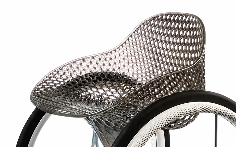 View of the middle section of a geometrical, 3D-printed chair seat from a 3D-printed prototype of a customized, futuristic-looking wheelchair, using multiple 3D printing materials. The seat is lattice-structured and made of a translucent gray resin. The wheel spokes are made of 3D-printed metal.