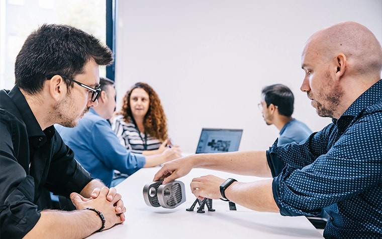 Two men sitting at a long table looking at metal 3D-printed parts with a group of three people talking around a laptop farther back at the table