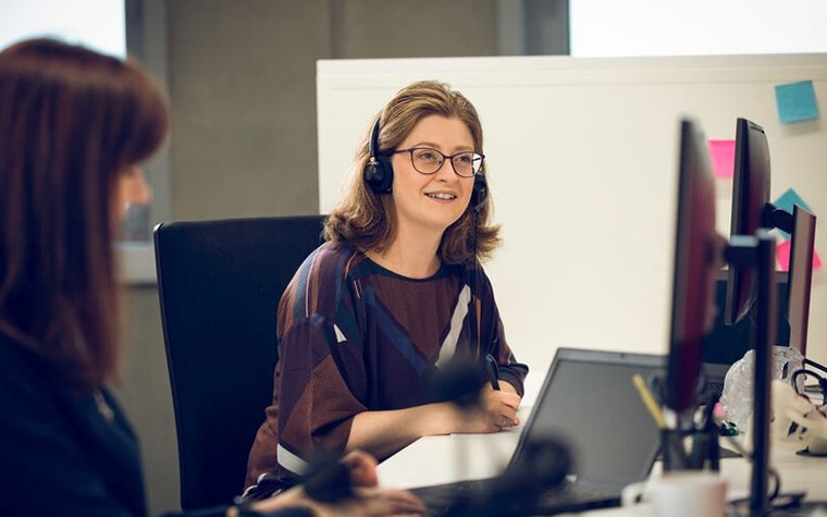 A woman with glasses sitting at a computer wearing a phone headset