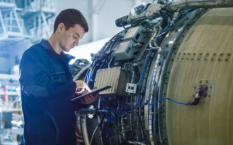 An engineer in a blue jumpsuit works on an electronic tablet next to a large airplane engine.