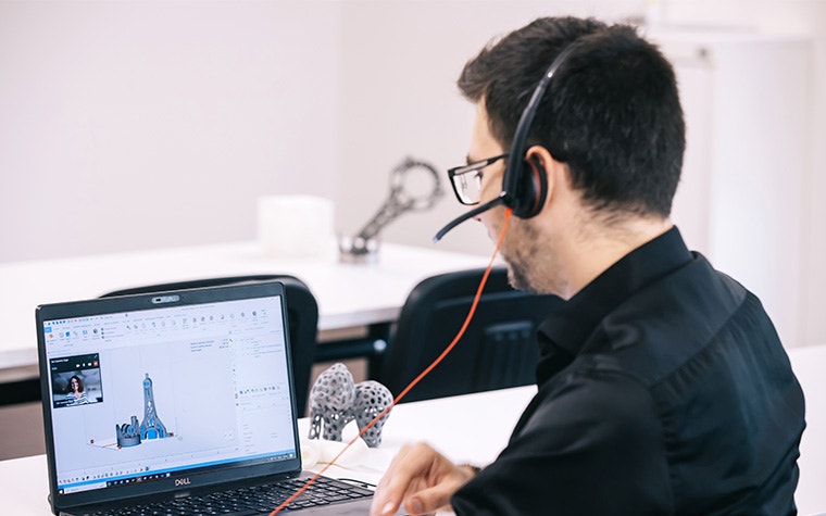 A man sitting at a table in front of a laptop with Magics pulled up and a small window with a Materialise Academy trainer giving a lesson. He's wearing a headset and there's a 3D-printed elephant on the table