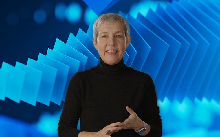 Die Vorstandsvorsitzende von Materialise, Brigitte de Vet, blickt in die Kamera vor einem Bildschirm mit blauen Quadraten, die von links nach rechts immer größer werden