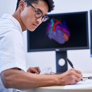 Man writing on a piece of paper at a desk in front of a computer showing medical software