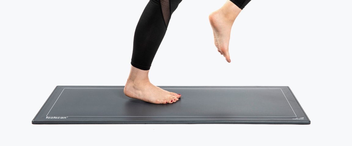 Eine Person steht auf einer Druckplatte, wobei der rechte Fuß flach auf dem Boden steht und das linke Bein leicht nach vorne angehoben ist