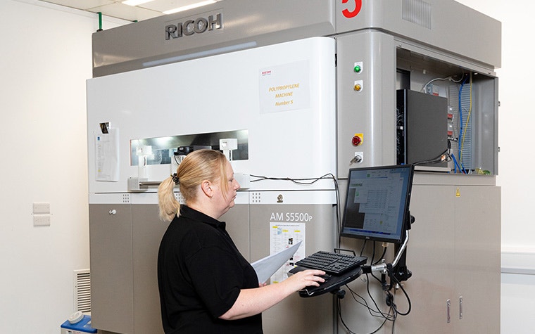 A Ricoh 3D production operator reviews a computer screen next to a 3D printer while holding a stack of papers