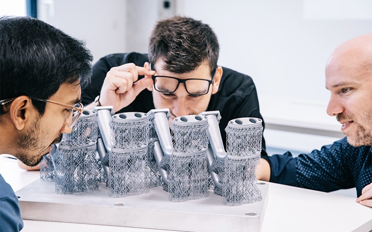 3 men looking closely at a metal 3D-printed build, including support structures, on a table