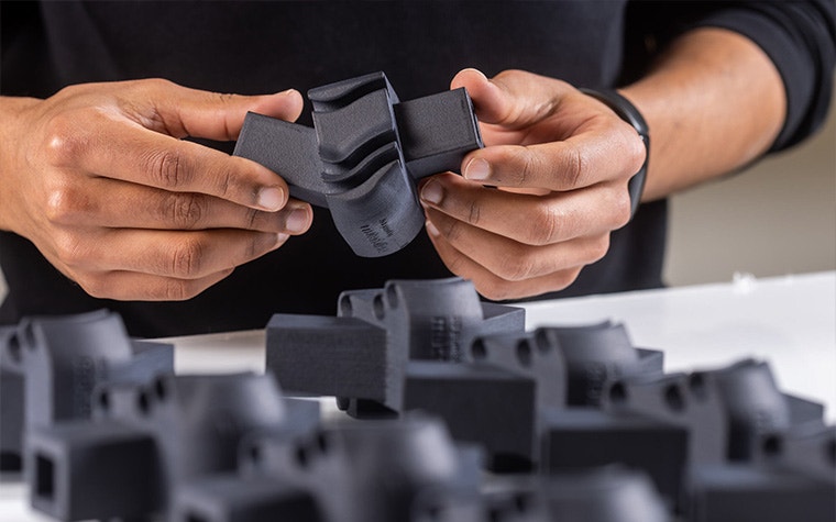 Hands holding one black 3D-printed part with copies of the same part on a white table