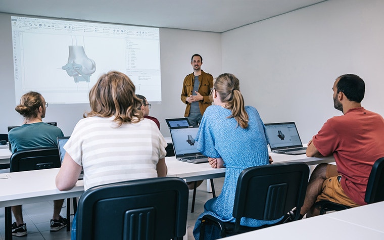 A Materialise Medical expert stands at the front of a classroom full of trainees on their laptops, teaching them about a 3D-printed surgical guide shown in Mimics software on a projector. The trainees are also looking at the same screen on their laptops.